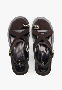 Hnědé semišové sandály s překříženými pásky a lesklým zlatým kovovým akcentem. Texturovaná černá stélka a světlá šedá podrážka dokončují design.