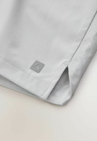 Gros plan sur l'ourlet d'un vêtement gris clair avec un petit patch logo triangulaire et une fente latérale révélant une doublure intérieure à pois.