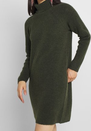Femme portant une robe pull à manches longues en tricot vert foncé avec un col roulé, debout, une main sur la hanche et l'autre détendue le long du corps.