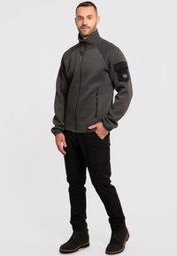 Szara polarowa kurtka na zamek z wysokim kołnierzem, bocznymi kieszeniami i czarną naszywką na rękawie. Noszona z czarnymi spodniami cargo i butami.