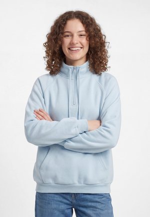 Giovane con capelli ricci sorride, indossa una felpa azzurro chiaro con zip a quarto e jeans blu, con le braccia incrociate davanti.