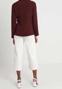 Blazer tailored borgogna con colletto alto, maniche lunghe e cuciture visibili; abbinato a pantaloni bianchi a gamba larga e sandali bianchi con tacco quadro.