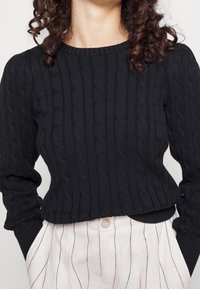 Černý pletený svetr s copánkovým vzorem, kulatým výstřihem a dlouhými mírně nařasenými rukávy. Krátký střih v pase.