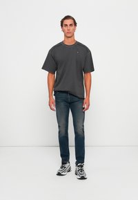 Male model stands against white wall wearing dark gray oversized T-shirt, faded blue jeans, and black-and-white athletic shoes.