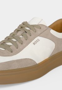 Μπεζ και λευκό παπούτσι BOSS με σουέτ λεπτομέρειες, ταμπά λαστιχένια σόλα, μπεζ κορδόνια και ορατό λογότυπο "BOSS" στο πλάι και στη γλώσσα.