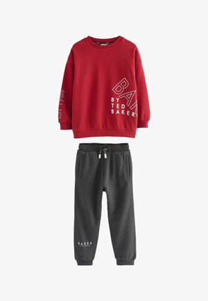 Młody chłopiec stoi, ubrany w czerwoną bluzę z długim rękawem z napisem "Baker by Ted Baker", ciemne spodnie i białe trampki z gumowymi podeszwami.