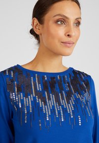 Maglione blu con scollo rotondo e un motivo di paillettes in nero, oro e argento che si sviluppa dalle spalle.