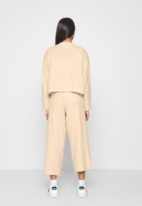 Maglione beige, corto, e pantaloni a gamba larga in materiale morbido e texturizzato. Abbigliamento abbinato a sneakers bianche, che mostrano un fit rilassato.