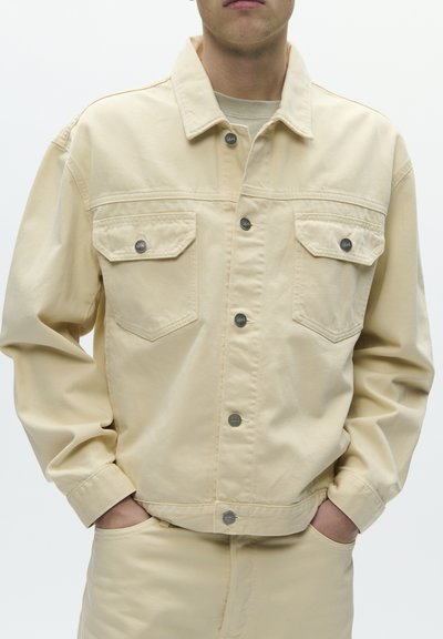 Man wearing beige denim jacket with buttoned chest pockets and matching beige pants, hands in pockets, against plain white background.