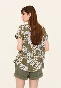 Camicia a maniche corte color verde oliva con un motivo floreale in blu chiaro e bianco, caratterizzata da una vestibilità ampia e un orlo curvo. Abbinata a pantaloni corti arrotolati.