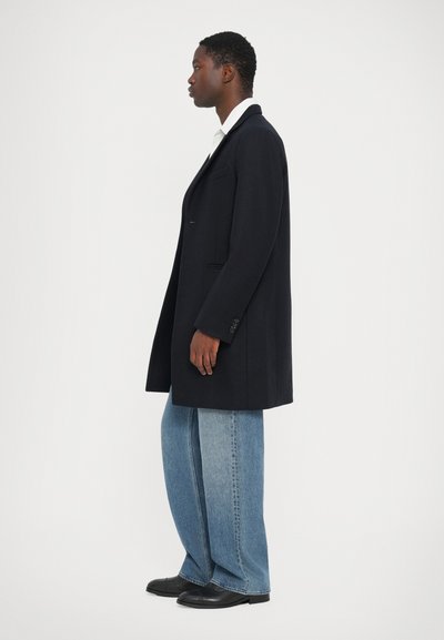 Cappotto in misto lana blu navy con una vestibilità su misura, bottone singolo e rever a punta, abbinato a jeans blu chiaro a gamba larga e scarpe nere.
