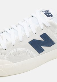 Sneakers ar baltu ādas un zamša virsu, zilu "N" logotipu, baltām auklām un teksturētu gumijas zoli. Iespraugas ar šuvēm.