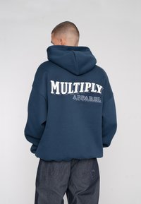 Navy blue hoodie with a large white embroidered "MULTIPLY APPAREL" on the back. Features a spacious hood and oversized fit.