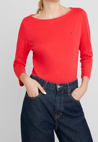 Kvinde iført en rød langærmet skjorte med et lille logo på brystet og mørkeblå højtaljede jeans med hænderne i lommerne.