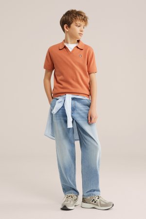 Garçon portant un polo orange à manches courtes, un jean bleu clair, des baskets grises et une chemise bleu clair nouée autour de la taille, debout et regardant vers la droite.