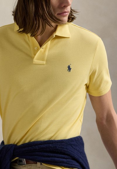 Muškarac u žutoj polo majici s malim tamnoplavim logom igrača polo, tamnoplavi džemper zavezan oko struka i hlače boje kakija.