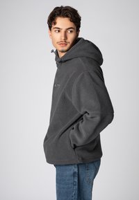Grauer Fleece-Hoodie mit Kapuze mit Kordelzug, Kängurutasche und strukturierter Oberfläche. Kombiniert mit blauen Jeans, vor einem hellen Hintergrund dargestellt.
