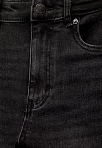 Gros plan d'un jean en denim noir montrant la ceinture, le bouton, la zone de la fermeture éclair, les passants de ceinture et la poche avant avec des détails en rivets métalliques.