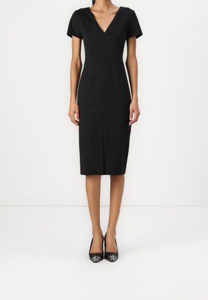 Vrouw draagt een nauwsluitende zwarte jurk tot op de knie met korte mouwen, een V-hals en zwarte pumps met spits toelopende neus, staand tegen een witte achtergrond.
