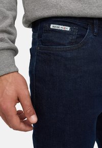 Gros plan sur la main d'une personne près de la poche avant d'un jean en denim bleu foncé avec une étiquette "INDICODE" visible.