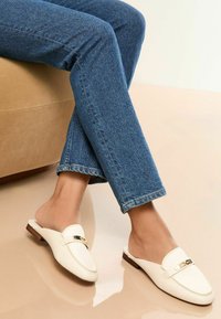 Witte leren instapschoenen met een gouden accent, platte hak en ronde neus, gedragen met blauwe denim jeans. Soepele textuur en minimalistisch design.