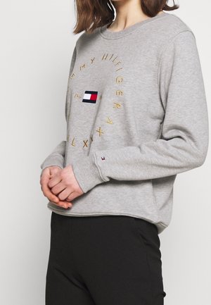 Persoon draagt een grijze Tommy Hilfiger-trui met gouden cirkelvormige tekst en logo op de borst, klein logo op de linkerpols, gecombineerd met zwarte broek.