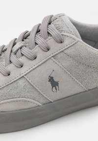 Grå mockaskor med en texturerad yta, platta grå snörningar och en svart logotyp av en ryttare på sidan. Gummisula.