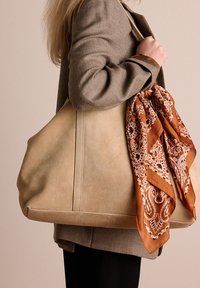 Beige suède tote bag met een ronde vorm, voorzien van een bruine patronen sjaal die aan de handvat is geknoopt, tegen een neutrale achtergrond.