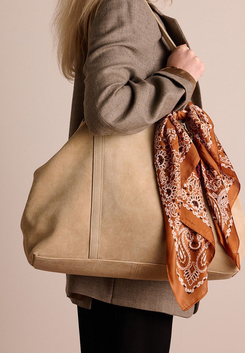Beige suède tote bag met een ronde vorm, voorzien van een bruine patronen sjaal die aan de handvat is geknoopt, tegen een neutrale achtergrond.