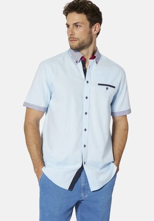 Mann trägt hellblaues, kurzärmeliges Hemd mit Knopfleiste, kontrastierendem Kragen und Taschenbesatz, kombiniert mit blauen Jeans, Hände in den Taschen.