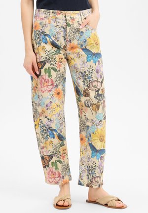 Broek met bloem- en vogelmotief in pastelkleuren, gedragen met beige sandalen, hand in de zak, tegen een effen achtergrond.