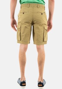 Pantaloni cargo beige con molte tasche, parte frontale piatta e tasca laterale con zip. Realizzati in tessuto leggero, abbinati a scarpe blu navy.