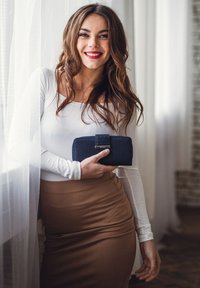 Navy wallet with a clasp and square pattern detail, held in a hand. Model wears a long-sleeve white top and a brown knee-length skirt.