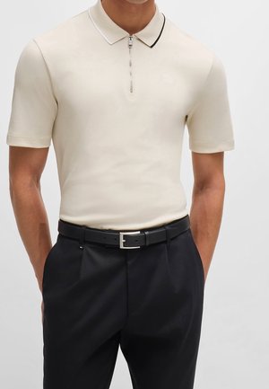 Beige Polo mit Reißverschluss am Hals und kurzen Ärmeln, mit einem kontrastierenden schwarz-weißen Kragen, kombiniert mit einer schwarzen Hose und einem schwarzen Gürtel.