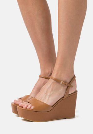 Brązowe sandały na koturnie z odkrytymi palcami i paskiem na kostce. Gładka skóra, krzywoliniowy kształt koturnu oraz platforma z przodu dla dodatkowego wzrostu.
