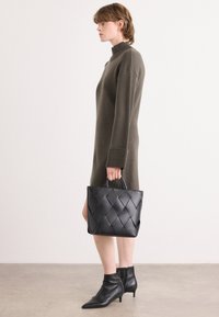 Μαύρη πλεκτή δερμάτινη τσάντα που κρατείται στο χέρι, συνδυασμένη με ένα ελαιοπράσινο, ριγέ φόρεμα με ψηλό λαιμό και μυτερές μποτάκια μέχρι τον αστράγαλο.
