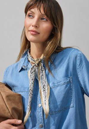 Žena nosí světle modrou džínovou košili a krémově bílý bandana s černým paisley vzorem uvázaný kolem krku, drží hnědou semišovou tašku.