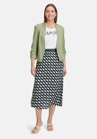 Veste vert clair texturée sur un t-shirt blanc avec écriture noire, associée à une jupe maxi à motifs navy et crème en vagues, et des sandales beiges.