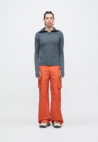 Top de manga comprida cinza claro com fecho de correr, combinado com calças cargo laranja vibrante. As calças apresentam bolsos laterais e um corte descontraído. Calçado casual completa o visual.