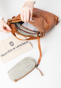 Bruine leren tas opengevouwen om een gestreepte binnenkant te onthullen; bevat ook een gestreepte pouch en een stoffen stofzak met een logo.