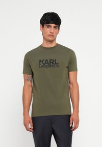 Olīvu zaļš īsām piedurknēm T-krekls, kas izgatavots no kokvilnas, ar apaļu kakla izgriezumu un melnu "KARL LAGERFELD" logo, kas drukāts krūšu daļā. Gluda tekstūra.