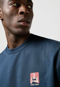 Mörkblå bomulls t-shirt med rund halsringning och ett grafiskt tryck av en karaktär i en röd och vit ruta på bröstet.