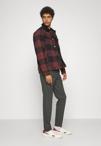 Chaqueta a cuadros roja y negra con cierre de botón, combinada con pantalones grises y zapatillas blancas con acentos rojos. El modelo está de lado.