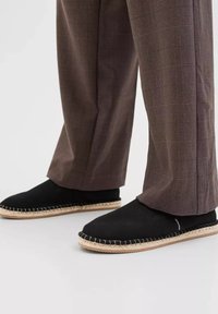 Black espadrilles with a woven jute sole and contrasting stitching, paired with plaid brown pants, showcasing a relaxed fit.