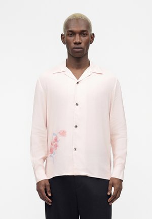 Άνδρας με ξανθά κοντά μαλλιά που φοράει μια ανοιχτό ροζ πουκαμίσα με κουμπιά και λουλουδάτο σχέδιο στο κάτω αριστερό μέρος, συνδυασμένη με μαύρο παντελόνι.