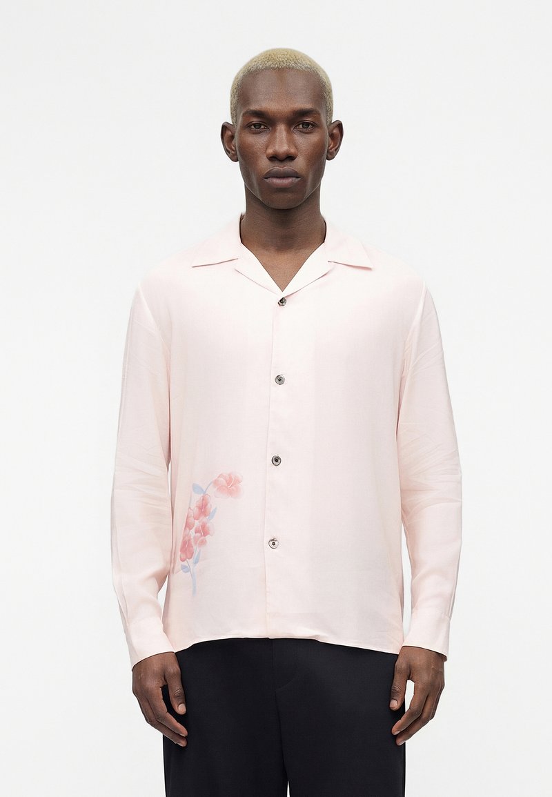 Άνδρας με ξανθά κοντά μαλλιά που φοράει μια ανοιχτό ροζ πουκαμίσα με κουμπιά και λουλουδάτο σχέδιο στο κάτω αριστερό μέρος, συνδυασμένη με μαύρο παντελόνι.