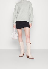 Beżowe kowbojskie buty z wysmukłym noskiem, falistymi krawędziami i obcasem na warstwach. Noszone z szarym swetrem i czarną mini spódnicą.