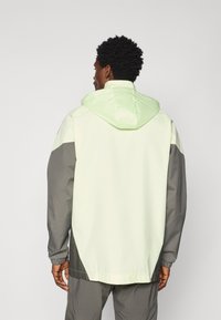 Lys grøn og grå vindjakke med hætte, der har elastiske manchetter og en buet kant. Glat tekstur, sporty design.