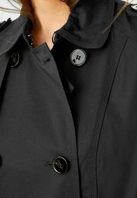 Trench-coat noir présentant un design croisé, de gros boutons et un col classique. Le tissu semble lisse avec un léger éclat.