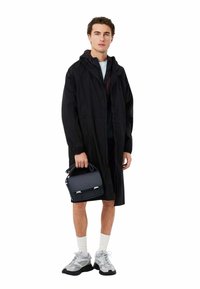 Cappotto lungo nero con cappuccio, indossato sopra un top scuro. Tiene una borsa blu navy con accessori in argento. Sneakers grigie con accenti neri. Calzini bianchi.